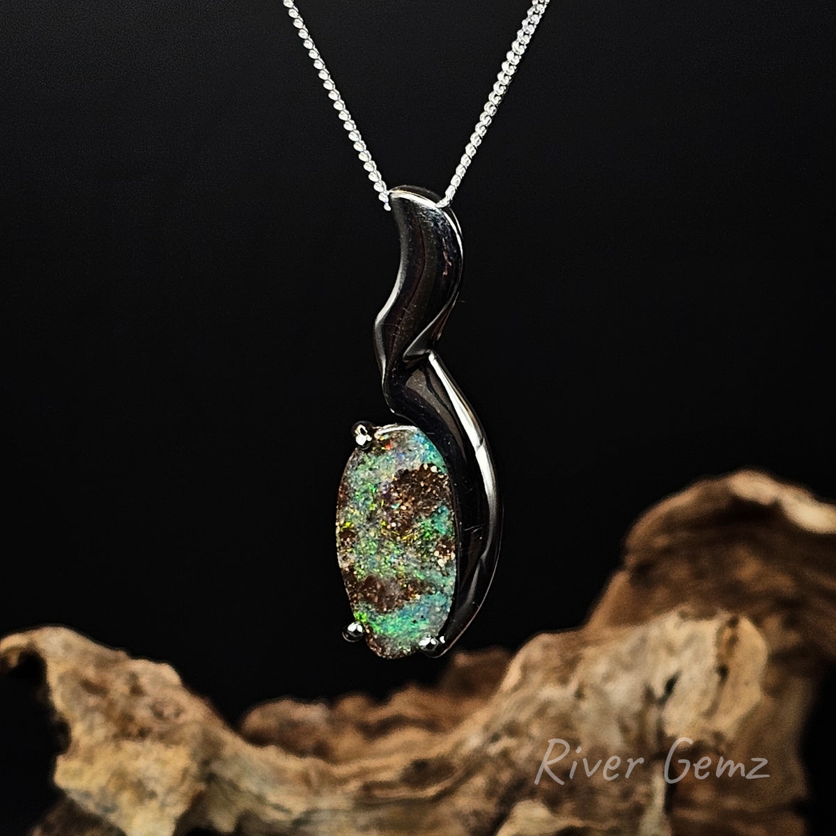 Green boulder opal besel and claw set in silver necklace against a black background with a drift wood base and 'River Gemz' water mark.