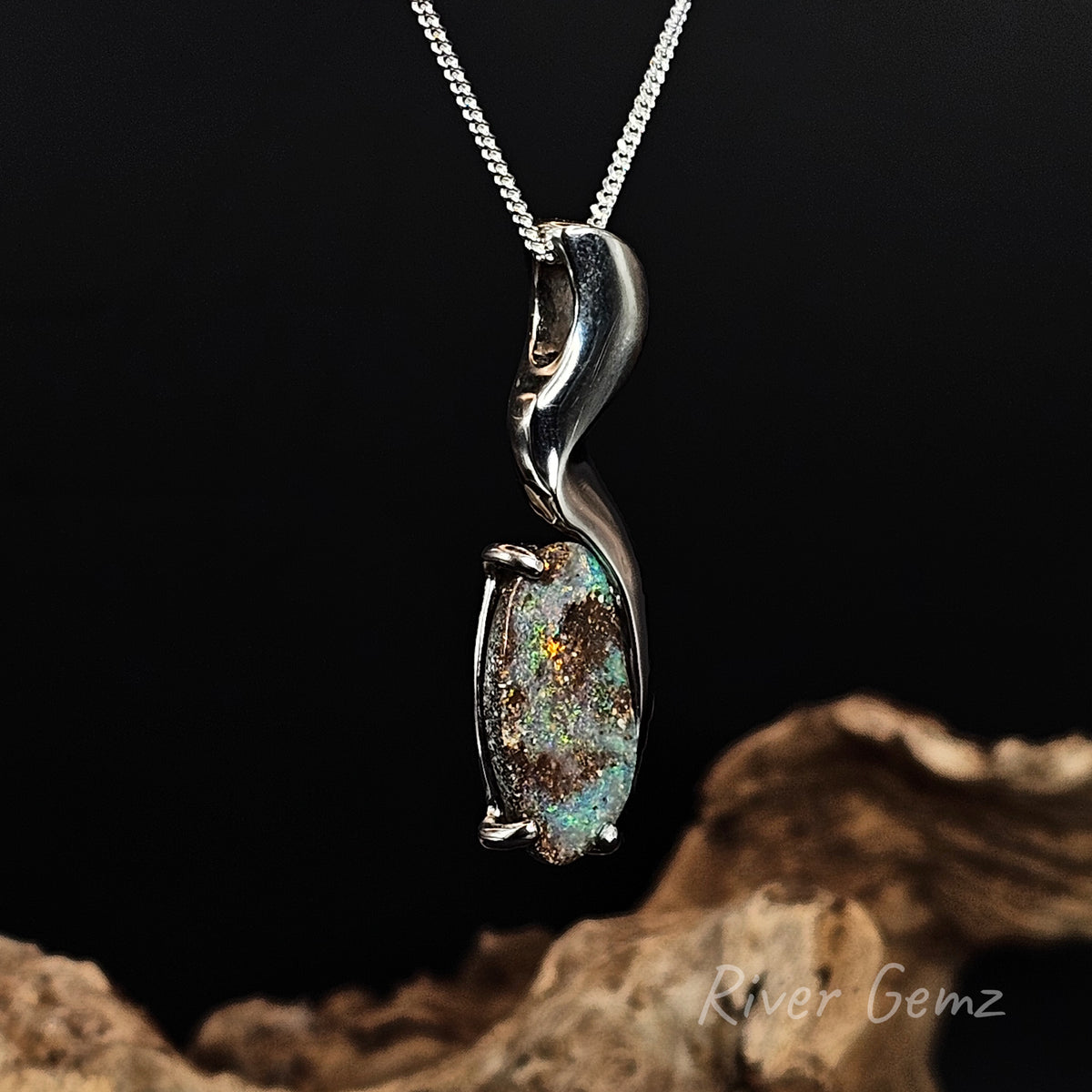 A silver pendant necklace featuring a curved, polished silver bail holding an oval iridescent opal gemstone with green, blue, and brown flashes, displayed against a black background with a piece of driftwood at the bottom.