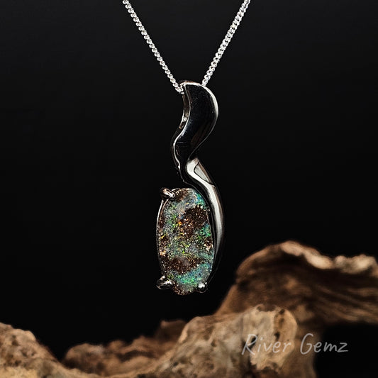 Oval shaped boulder opal with green colouring on a black background and piece of drift wood at the base with 'River Gemz' branding.