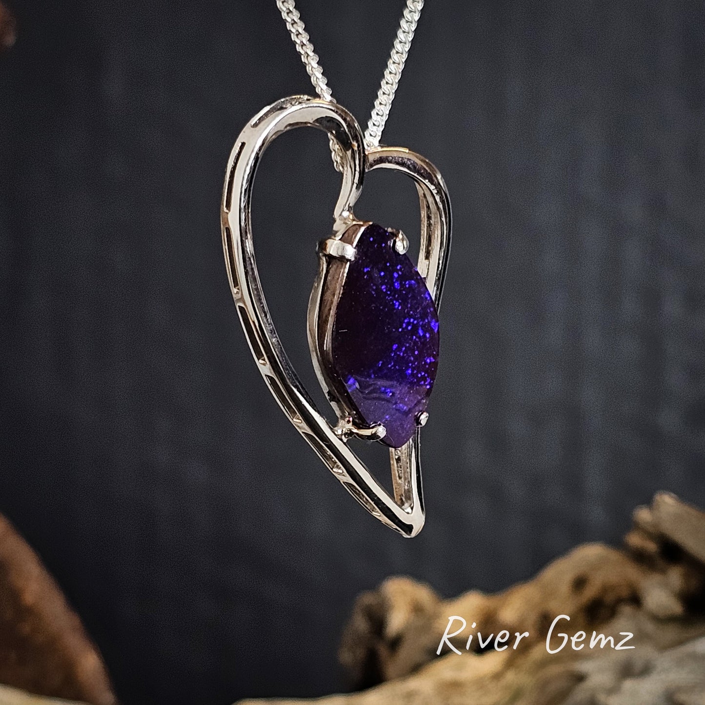 Silver heart-shaped necklace with a diamond shaped purple boulder opal on a dark background, featuring 'River Gemz' branding.