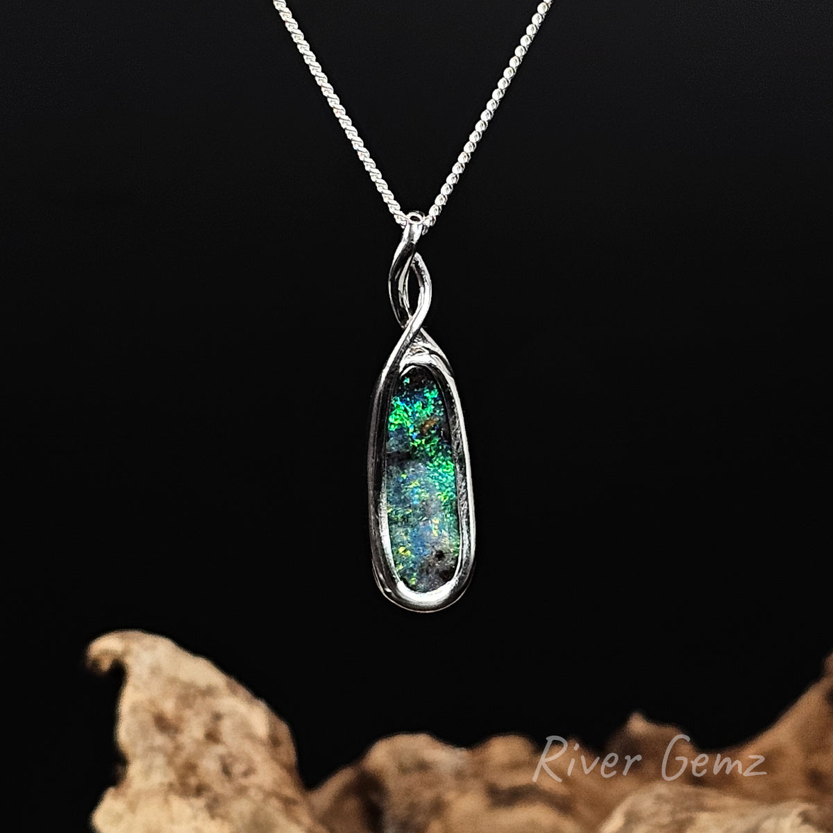 A silver pendant necklace featuring a oval-shaped opal gemstone with vibrant green, blue, and black play-of-color, displayed on a dark background with a piece of driftwood at the bottom. The brand name 'River Gemz' is visible in the bottom right corner.