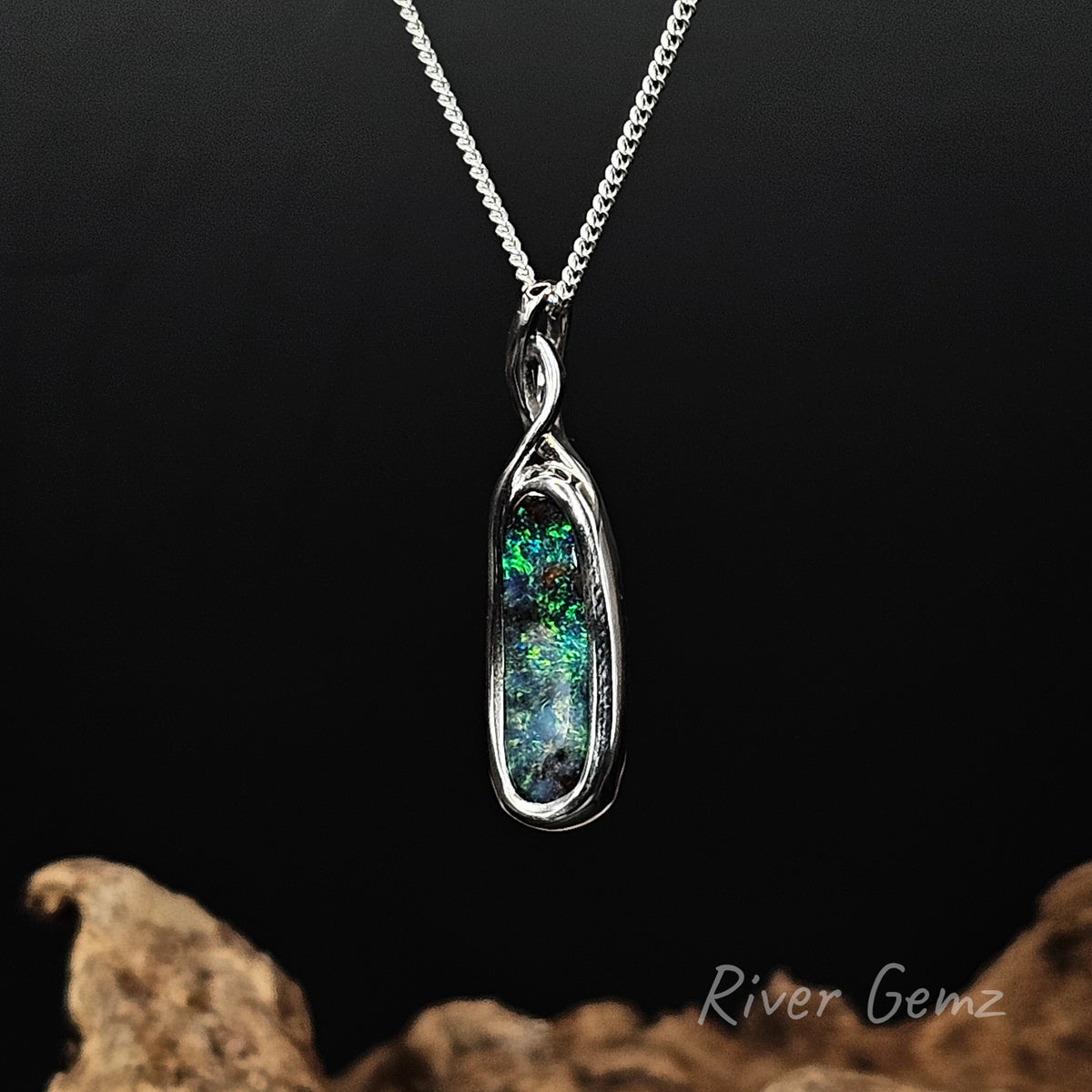 A silver chain necklace with a vertically oriented oval pendant featuring a vibrant green and blue iridescent opal in a bezel setting, displayed against a dark background with decorative driftwood at the bottom.