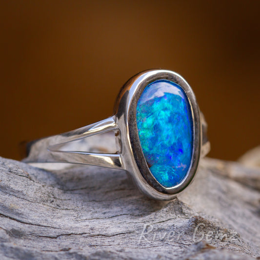 Approximately oval shaped blue-green opal in a silver ring. The ring is displayed on a silver-grey coloured bit of wood and has an orange/brown backdrop.