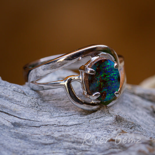 Vivid blue green speckles of colour throughout the boulder opal which is secured with 4 claws in the silver ring. Ring is shown on a grey weathered piece of driftwood and has a fire orange background.