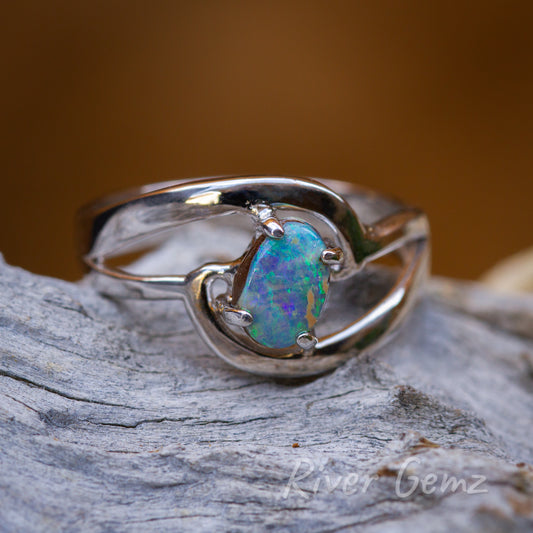 Free-form cut approximately oval shaped opal held securely with 4 claws in the silver ring. Ring is pictured on a weathered grey piece of wood and a sun fire orange back ground.