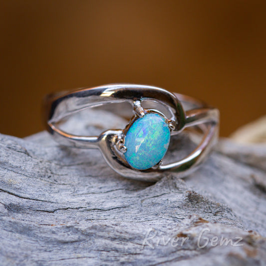 Ocean coloured oval shaped opal held securely by 4 claws in the silver ring. Ring is on a weathered grey piece of driftwood with an orange/brown background.