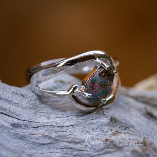 Free form cut multi-coloured opal secured with 4 claws in the silver ring. Ring is on a light-grey textured wood and pictured with a burnt orange back ground.