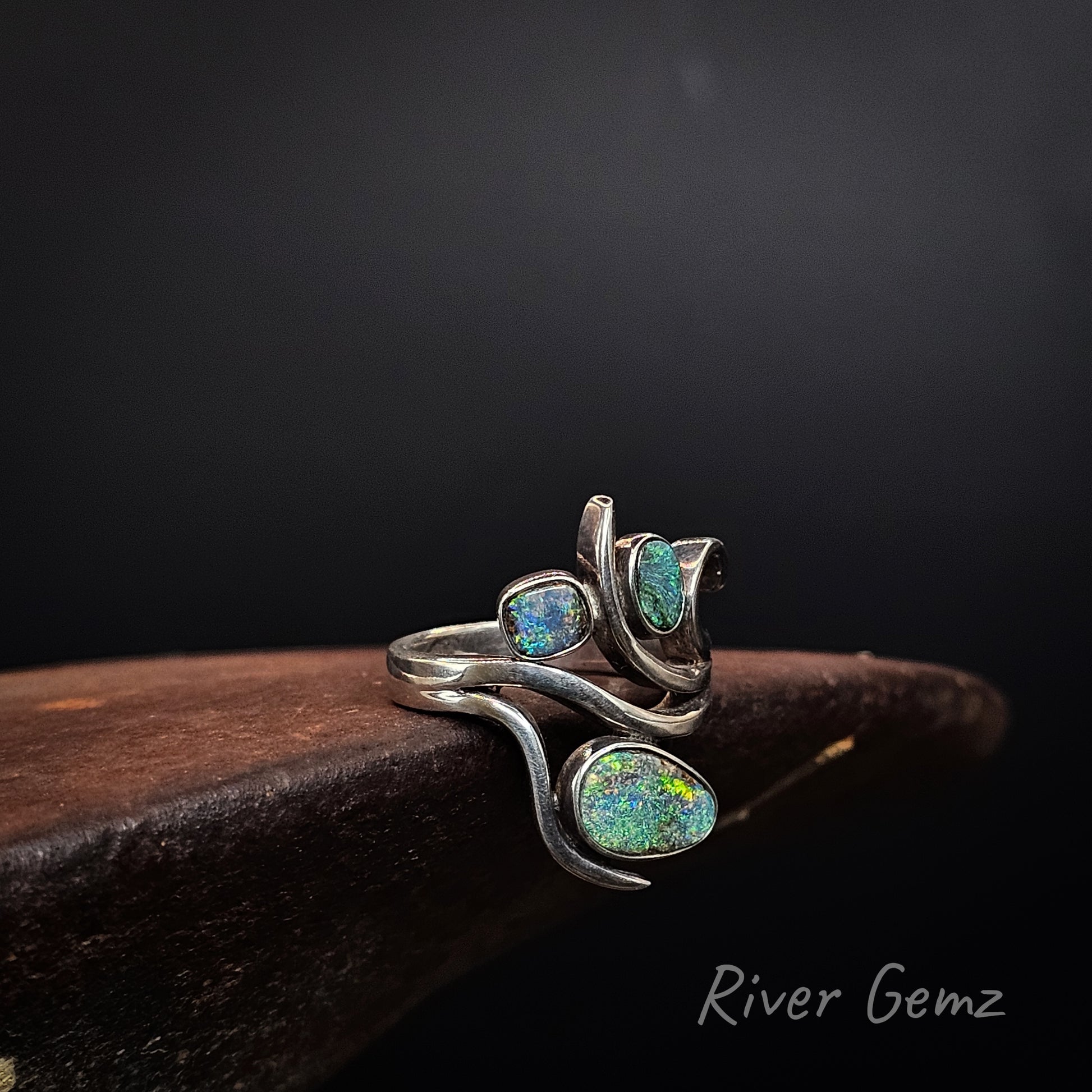 A silver ring featuring three oval green opal gemstones with iridescent blue and yellow flashes, displayed on a dark wooden block against a black background. The brand name 'River Gemz' is visible in the bottom right corner.