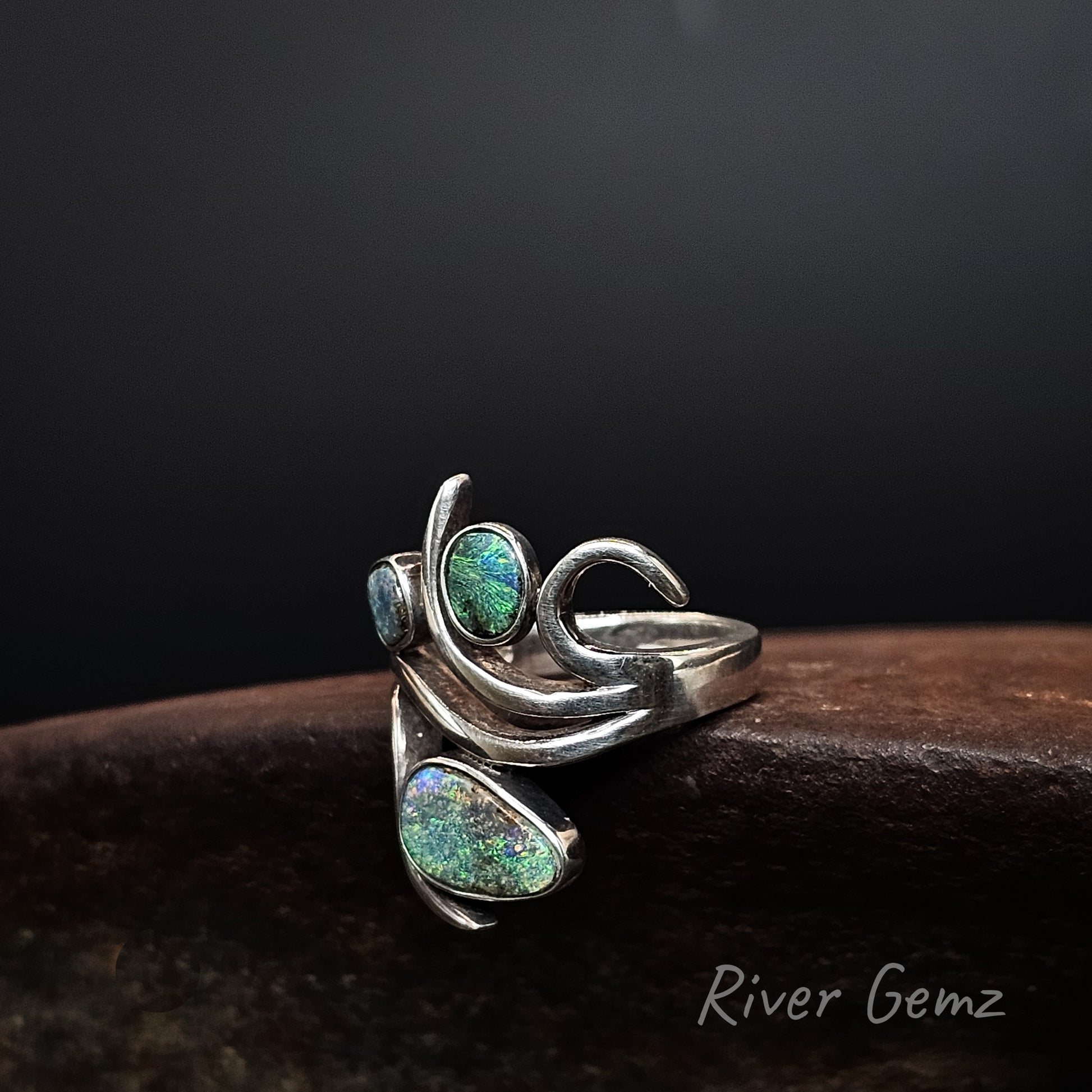 A silver ring with three iridescent green-blue opal stones, displayed on a dark wooden surface against a black background. The brand name 'River Gemz' is visible in the bottom right corner.