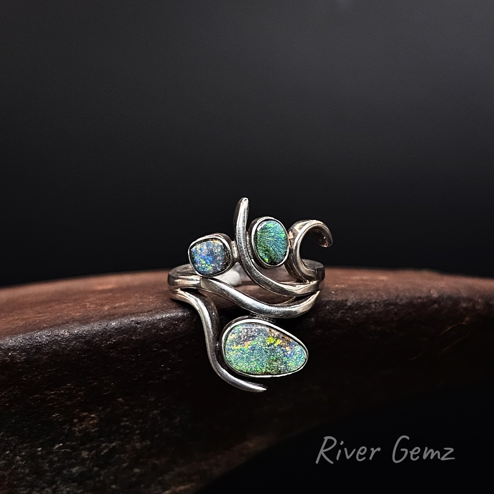 A silver wrap ring featuring three iridescent opal gemstones in blue, green, and multicolor hues, displayed on a dark wooden surface against a black background. The brand name 'River Gemz' is visible in the bottom right corner.