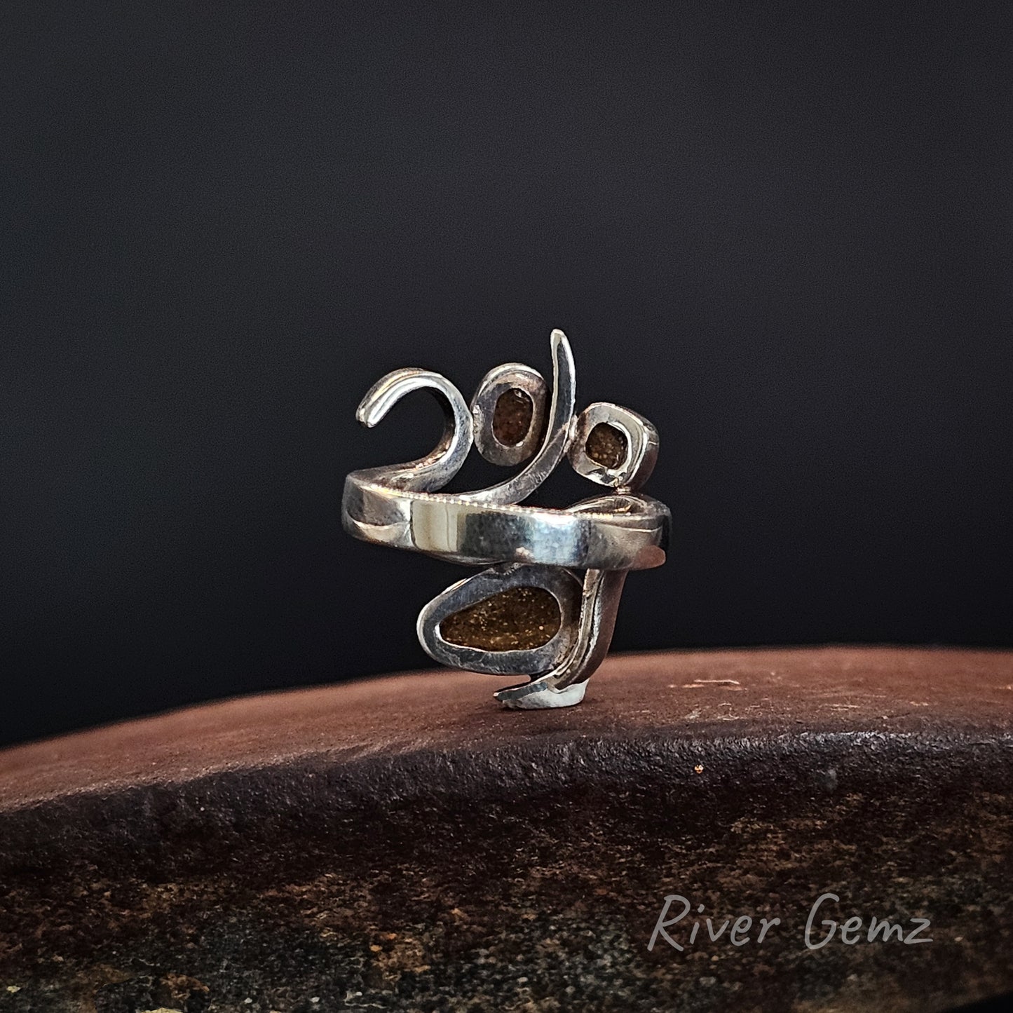A sterling silver ring shaped like the year 2020, featuring textured, polished silver bands and inlaid brown gemstone accents, displayed on a dark wooden surface against a dark background with a 'River Gemz' watermark.