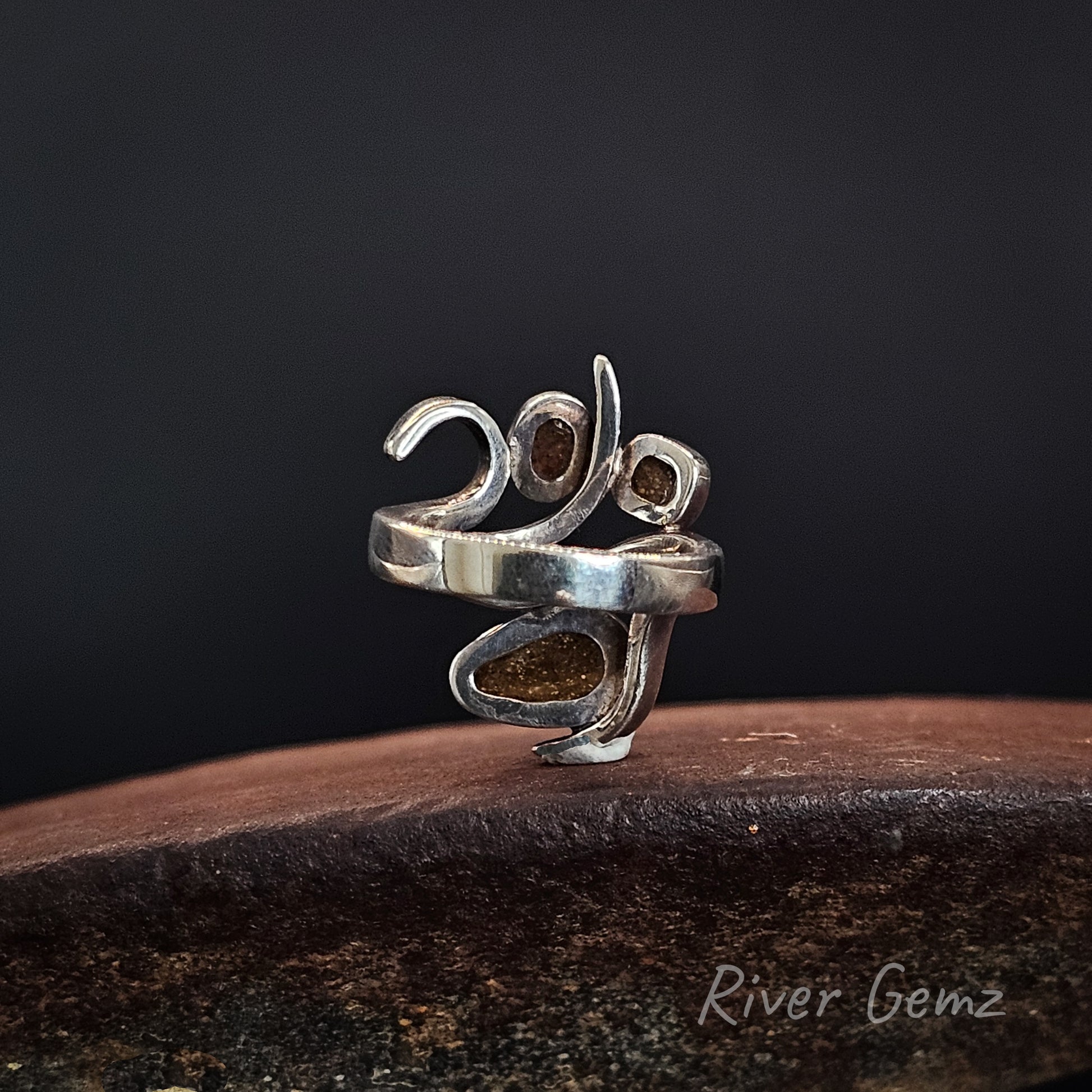 A sterling silver ring shaped like the year 2020, featuring textured, polished silver bands and inlaid brown gemstone accents, displayed on a dark wooden surface against a dark background with a 'River Gemz' watermark.