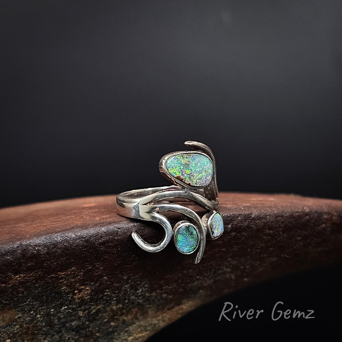 A sterling silver ring with three iridescent green-blue opal stones, displayed on a dark wooden block against a black background. The brand name 'River Gemz' is visible in the bottom right corner.