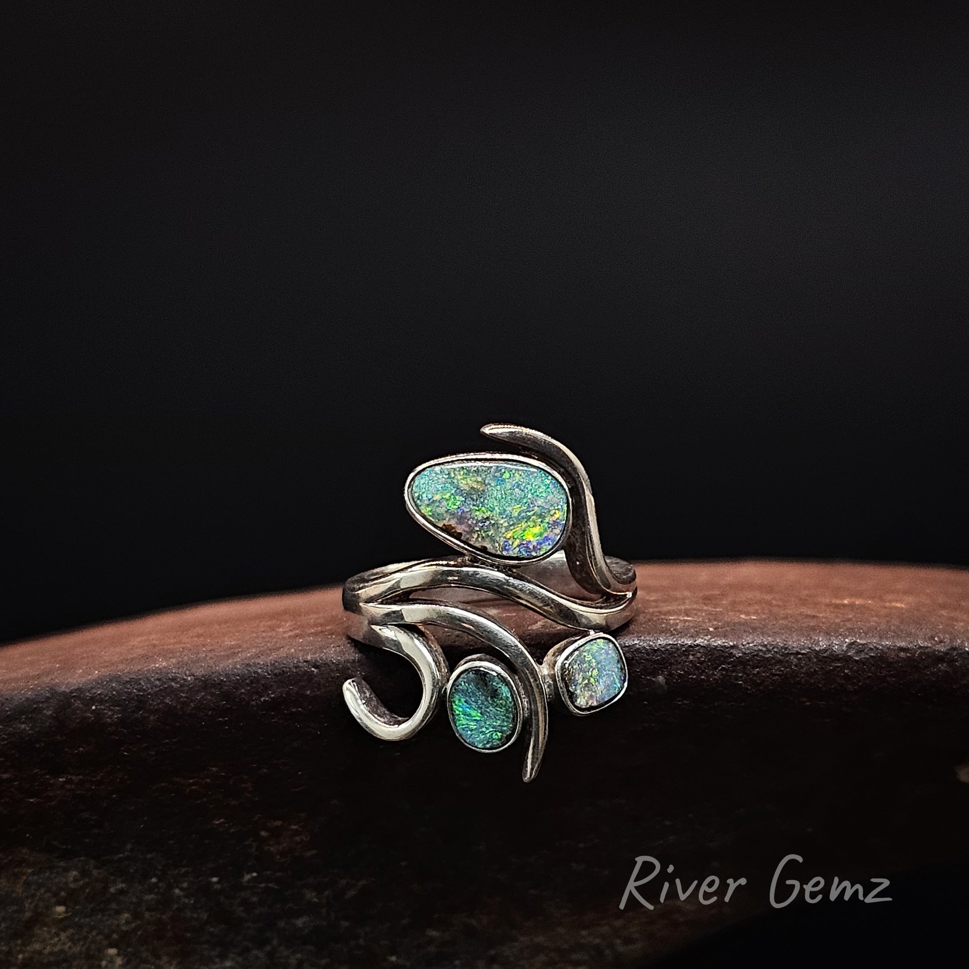 A silver wrap ring featuring three iridescent opal gemstones in organic shapes, displayed on a dark wooden surface against a black background. The brand name 'River Gemz' is visible in the bottom right corner.