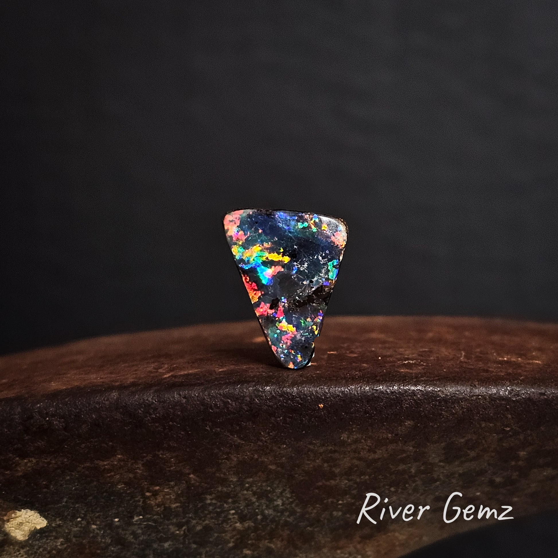 Multicolored triangular black boulder opal on a dark background with 'River Gemz' branding.