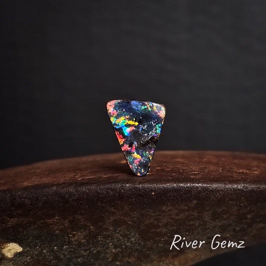 Multicolored triangular black boulder opal on a dark background with 'River Gemz' branding.