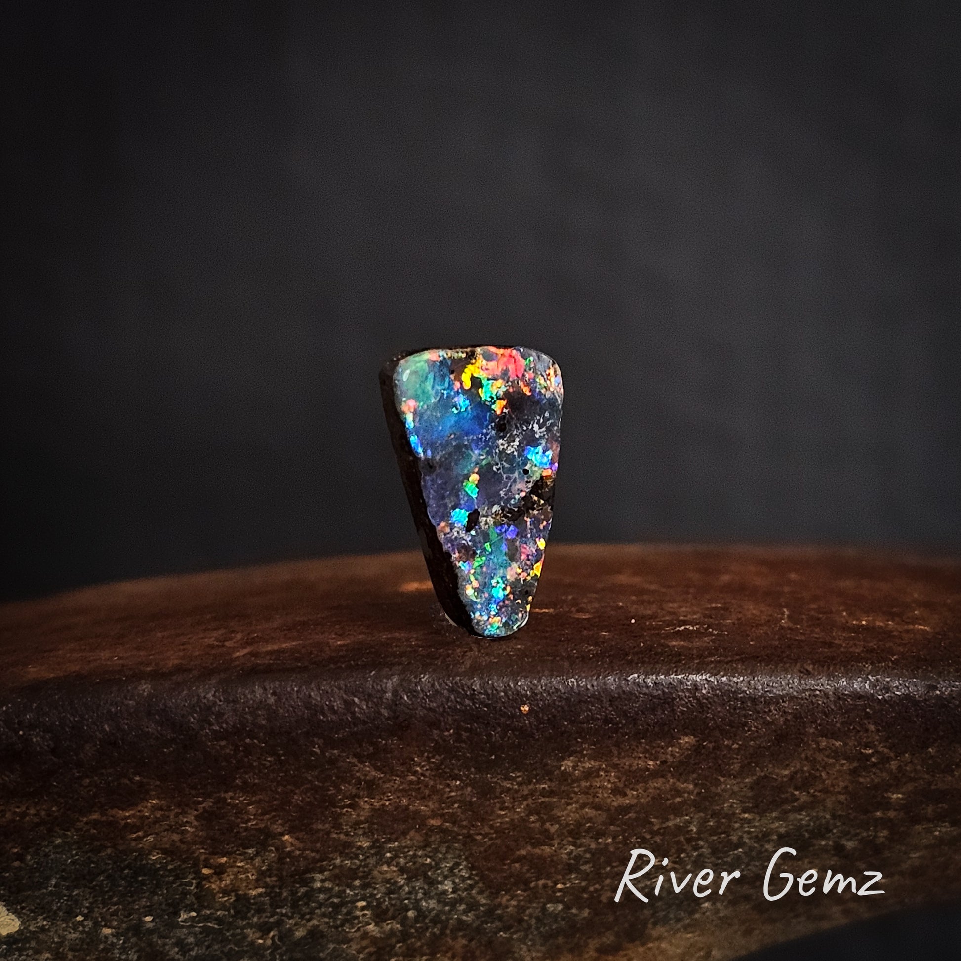 Multicolored unset dark opal on a dark background with 'River Gemz' branding.