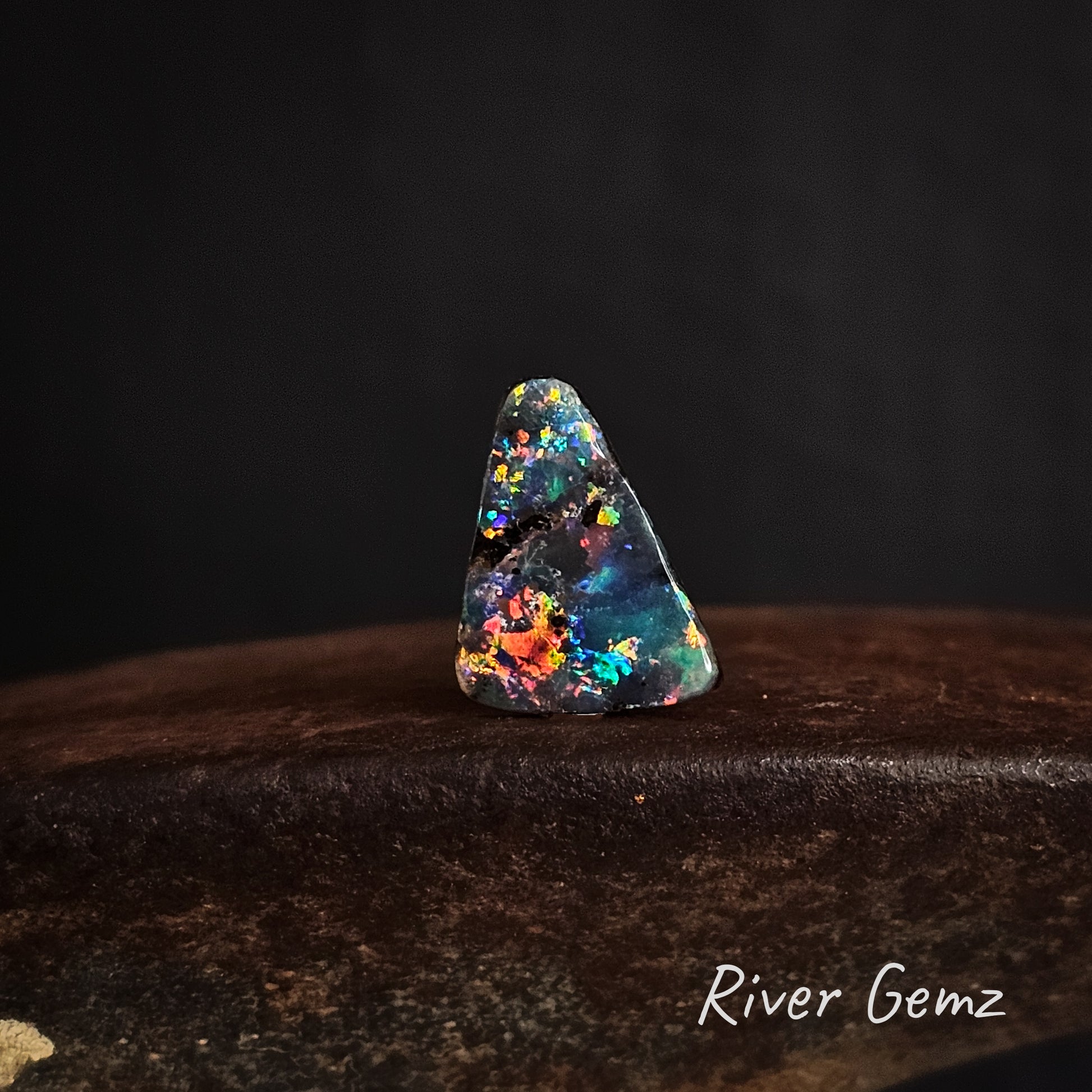 Multicolored triangular shaped opal stone on a dark background with 'River Gemz' branding.