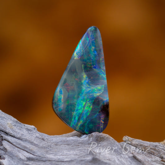 Front view of free form triangular shaped natural opal. Opal is sitting on a light grey weathered piece of drift wood with burnt orange back drop.