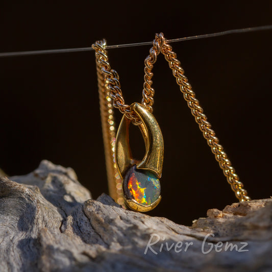 Orange, red flame on the left of the boulder opal and flashes of green on the right side. The opal is partial-besel set in the pendant. The bail is large and an integrated part of the design. Gold contrasts with dark backdrop and the light-grey grained driftwood.