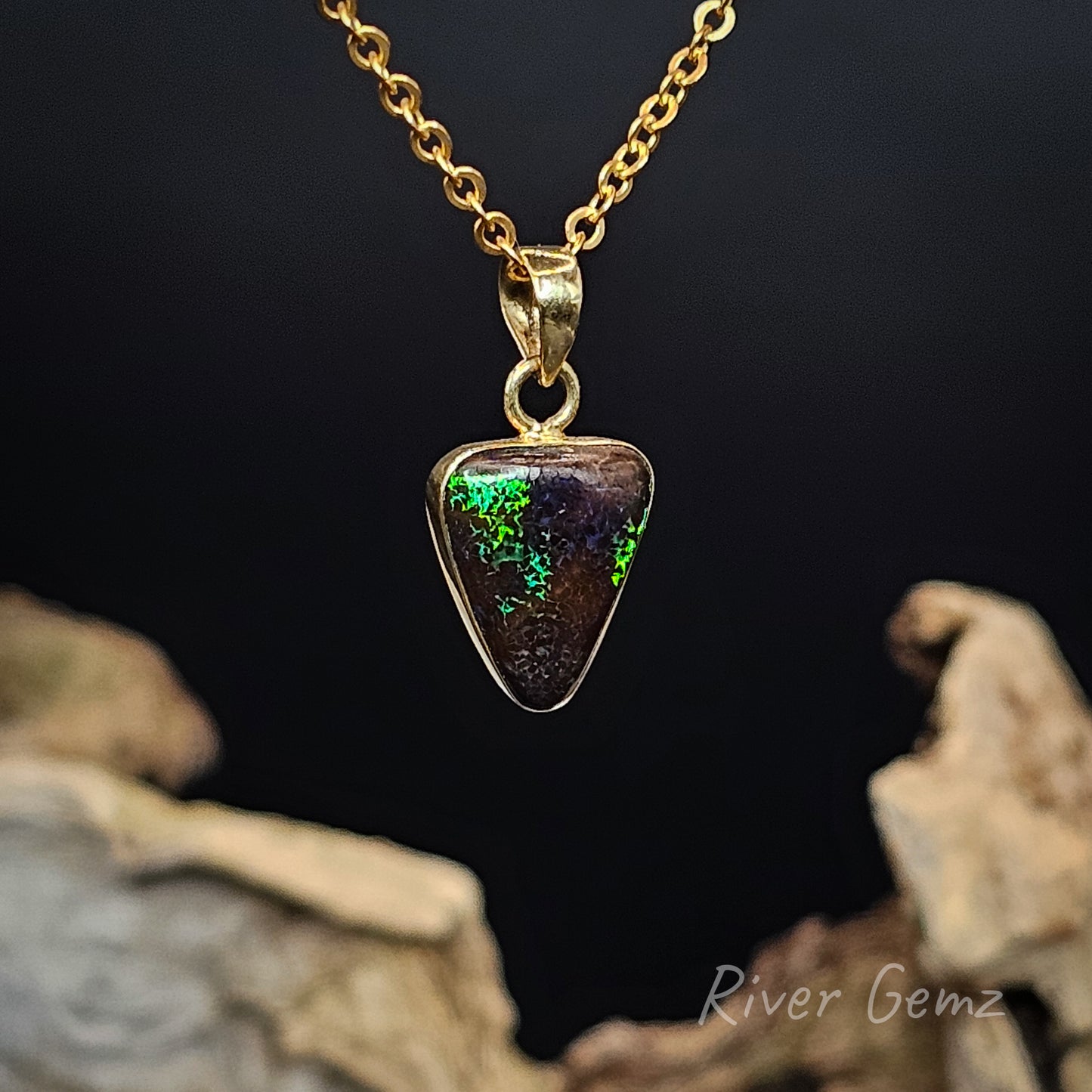 Gold necklace with a triangular pendant featuring green and purple colours, set against a dark background.