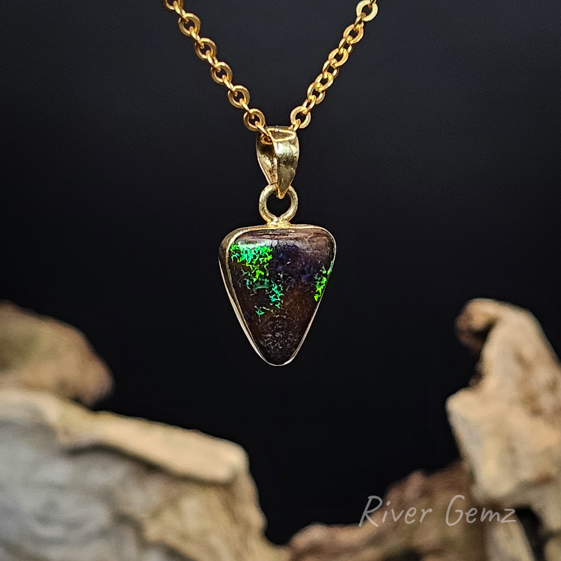 Gold necklace with a triangular pendant featuring green and purple colours, set against a dark background.