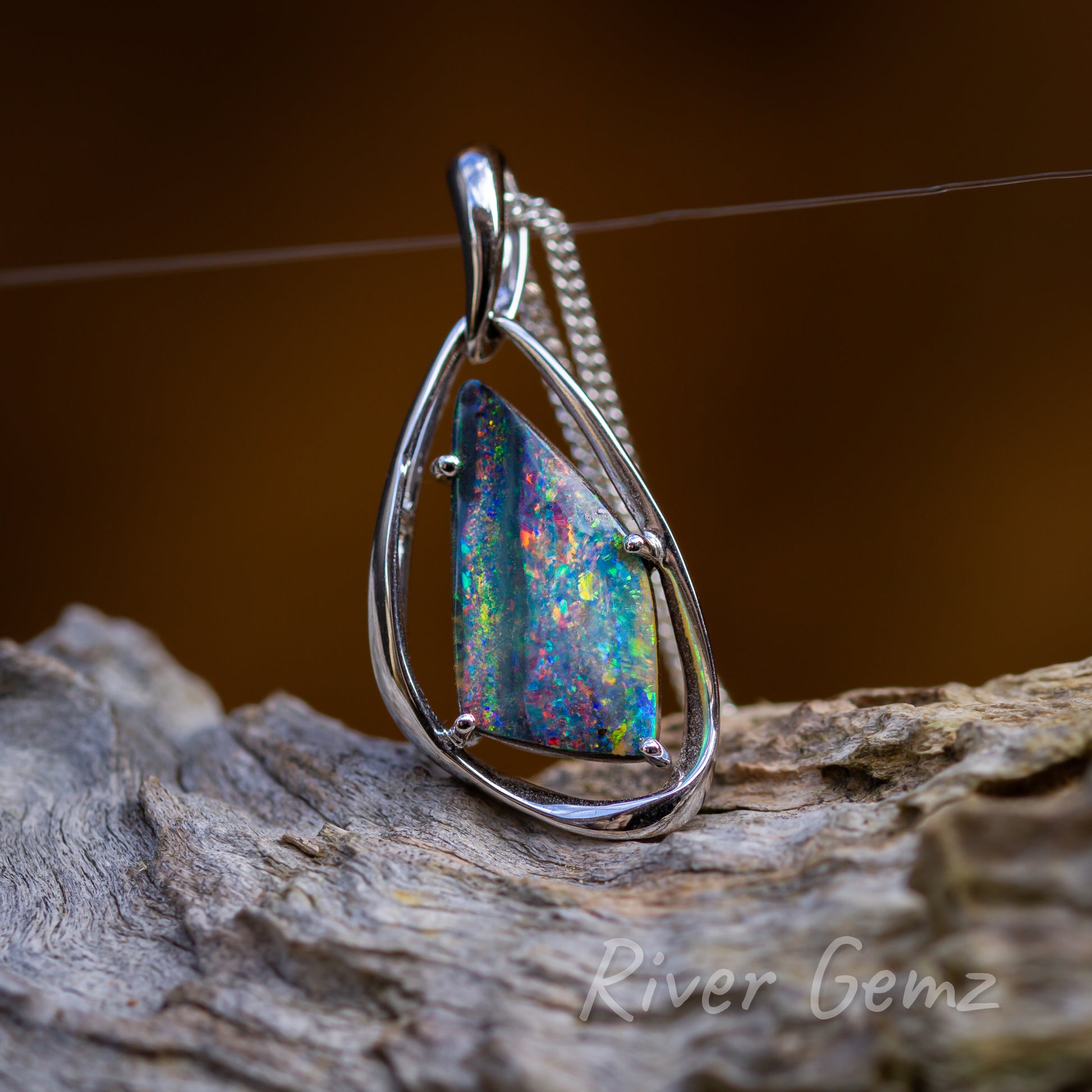 Free form cut multi-coloured opal with four sides held securely with 4 claws in the white gold setting including a stylish bail. Pendant is resting above a light-grey piece of driftwood and has a burnt orange backdrop.