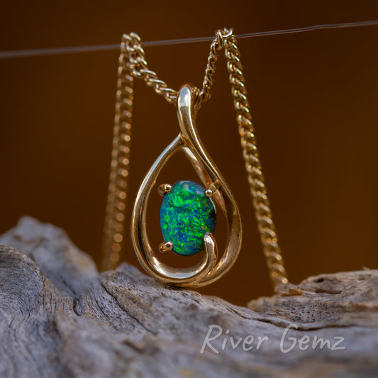 The blue-green colours are clearly evident in the opal secured by 4 claws to the rain drop shaped pendant.  The chain passes through a hole created in the setting and hence no bail is needed. The chain is hanging over a fishing line to display the opal upright. Gold contrasts well with the light-grey drift wood which the pendant is resting above.