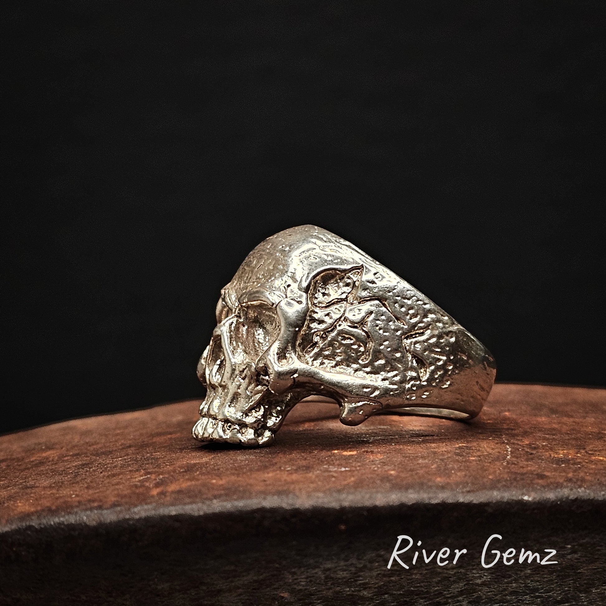 Side view of silver skull ring on a cast iron surface with a dark background, branded 'River Gemz'.