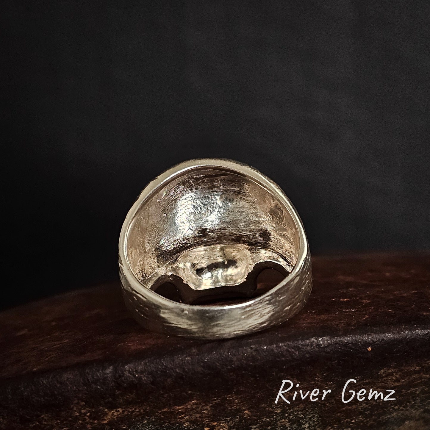 Rear view of silver ring on a dark brown surface with 'River Gemz' branding.