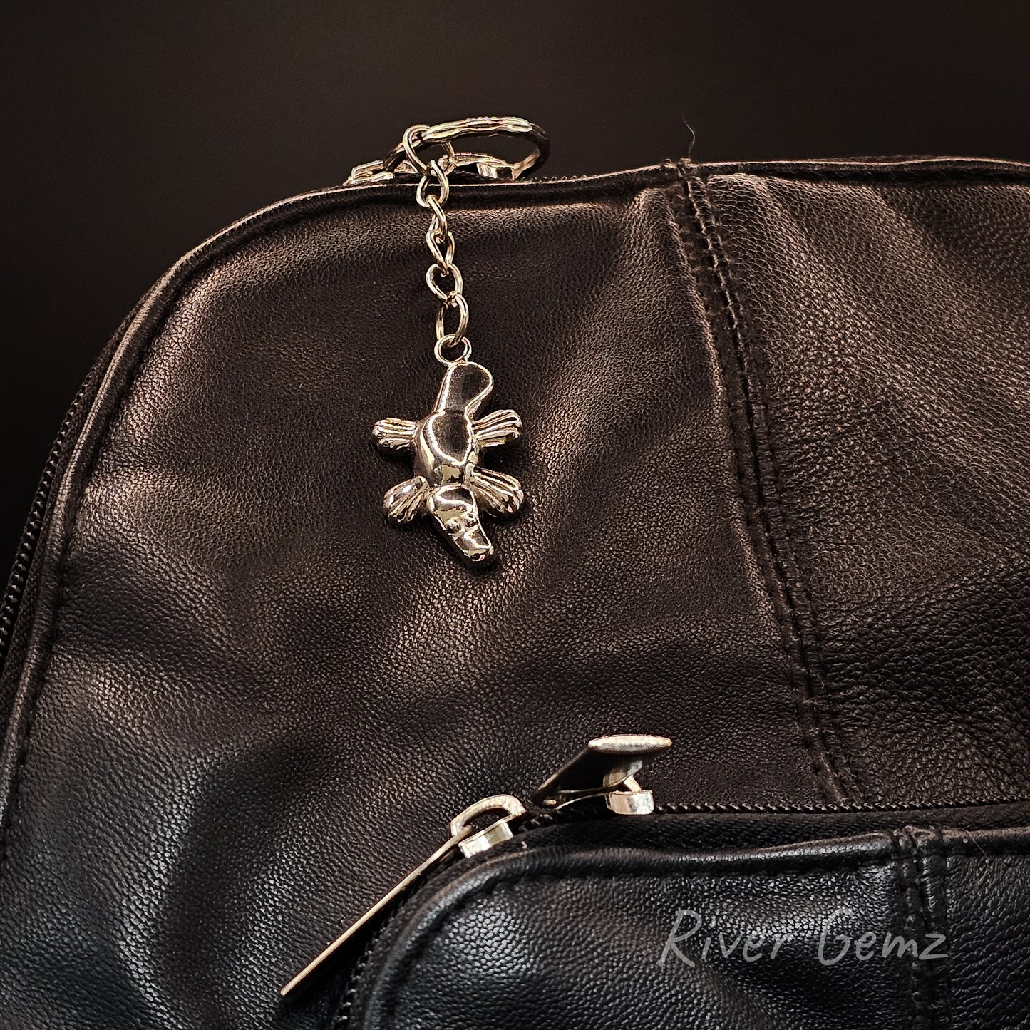 A close-up of a black leather bag featuring a silver metal zipper pull with a platypus-shaped charm, and a lower zipper with a silver pull. The brand name 'River Gemz' is printed in white script on the bottom right.
