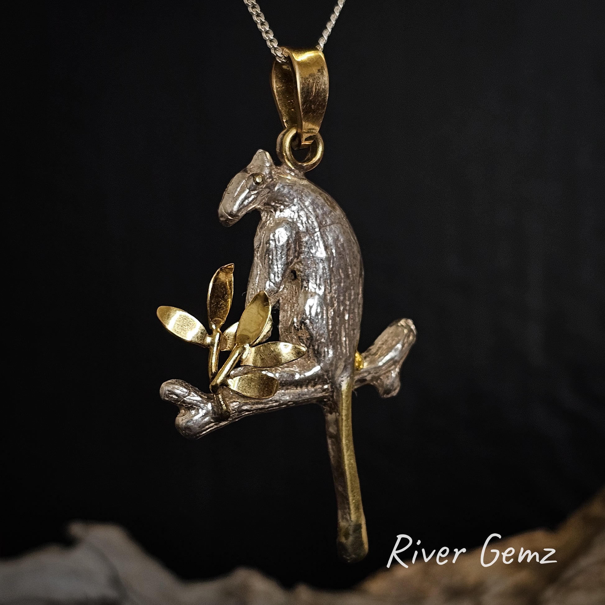 Gold and silver pendant of a tree kangaroo on a branch with leaves on a dark background, branded 'River Gemz'.