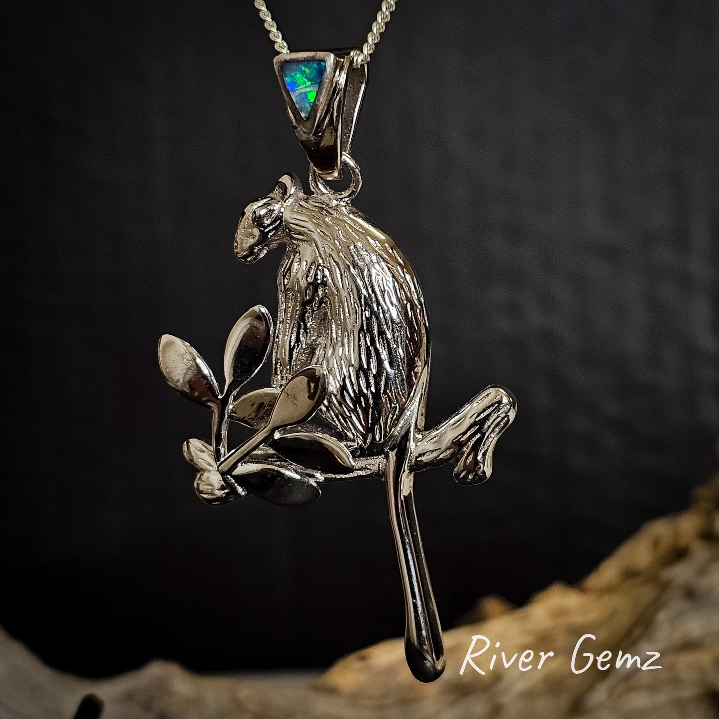 Silver pendant of a tree kangaroo on a branch with a dark background, featuring 'River Gemz' branding.