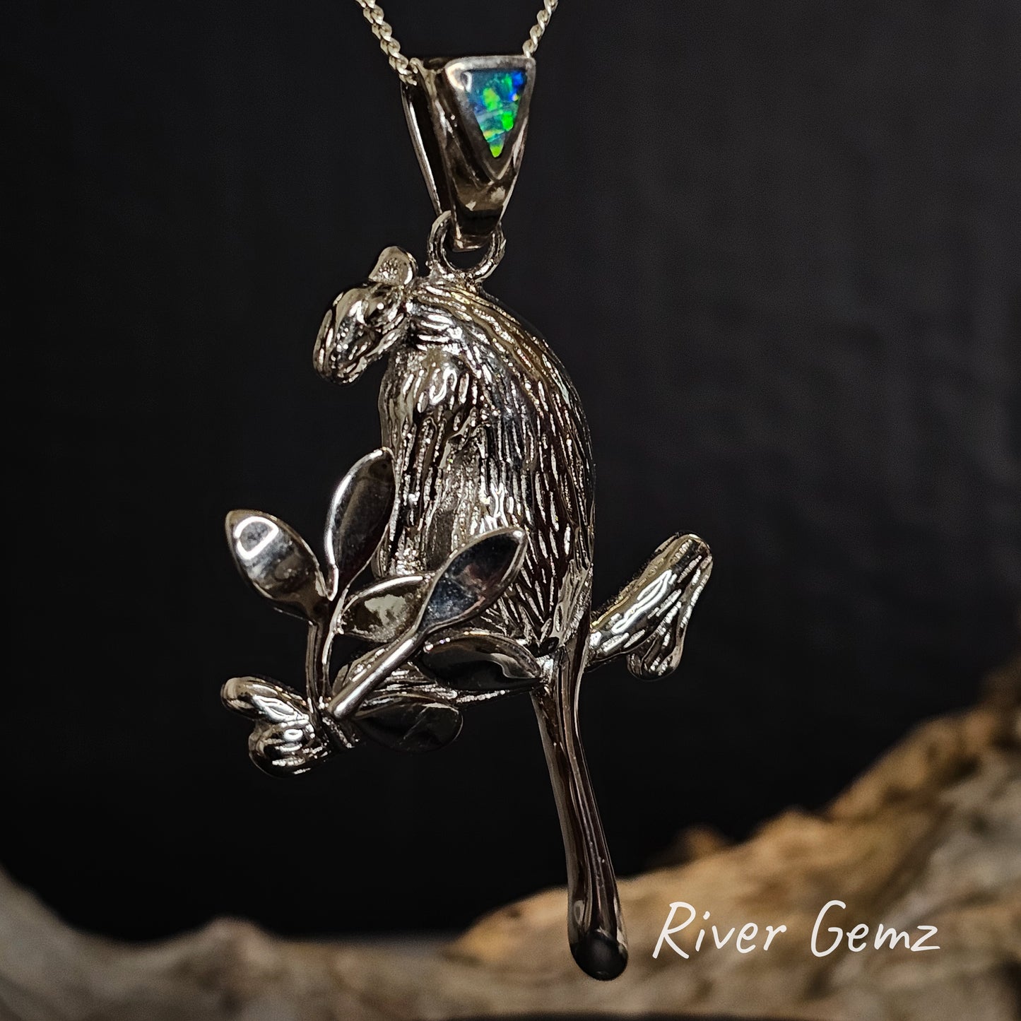 Silver tree kangaroo pendant with a blue and green opal in the bail on a dark background, branded 'River Gemz'.