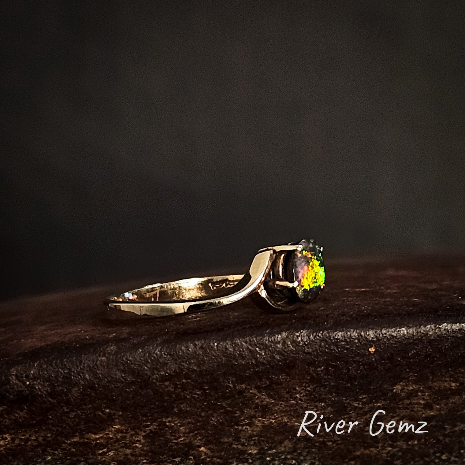 Side view of yellow gold ring with a multi-coloured dark opal on a dark surface, branded 'River Gemz'.