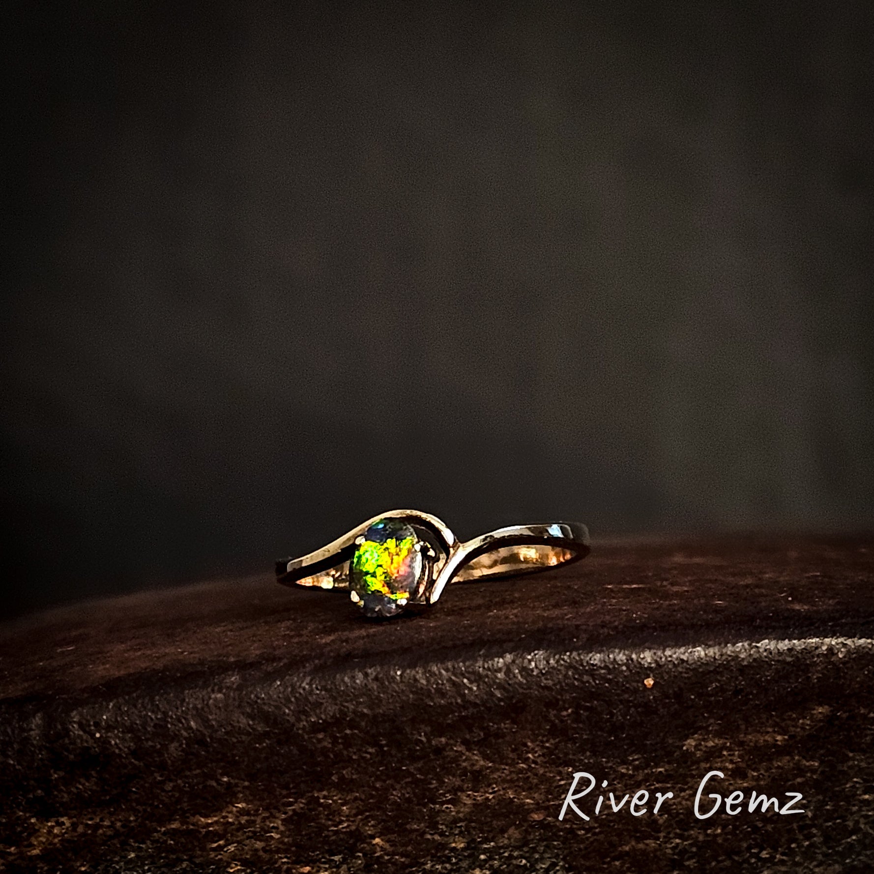 Multi-coloured dark opal claw set in narrow banded yellow gold ring show on cast iron surface branded 'River Gemz'.