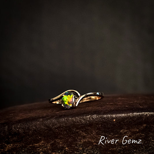 Multi-coloured dark opal claw set in narrow banded yellow gold ring show on cast iron surface branded 'River Gemz'.