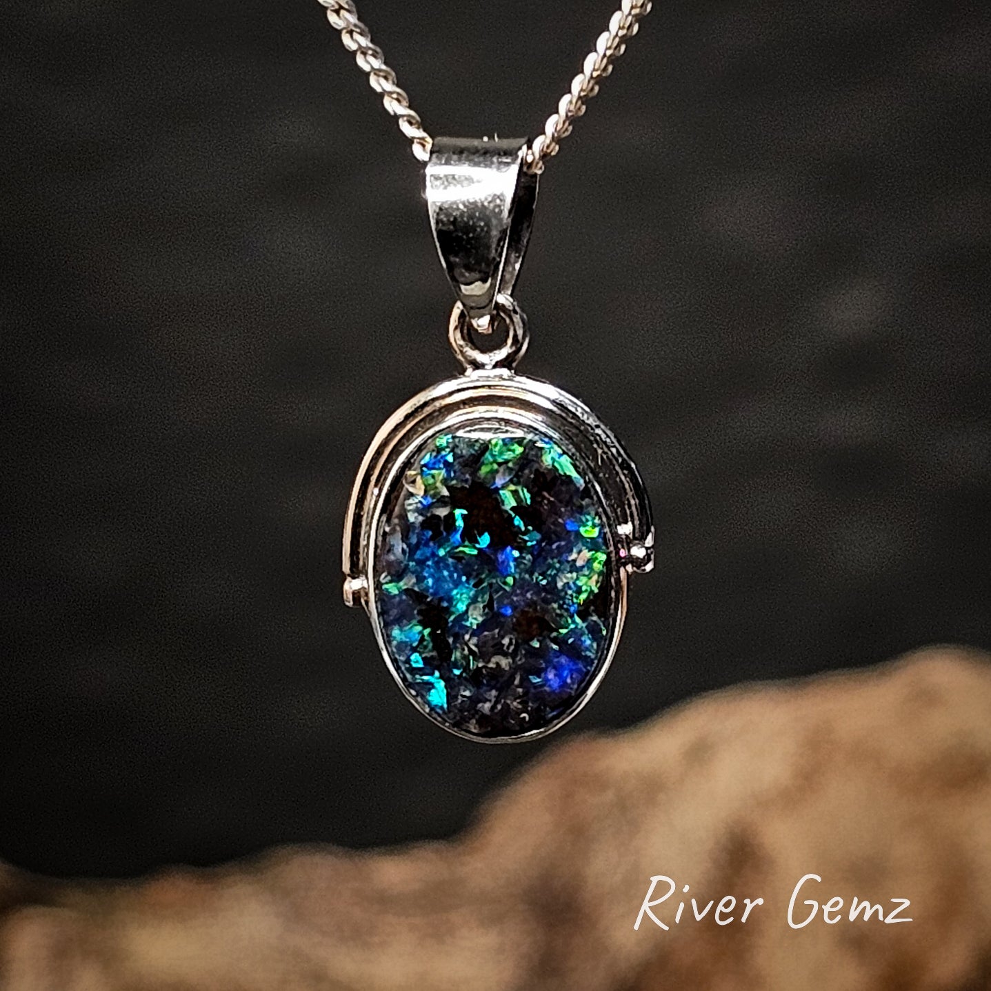 White gold pendant with an oval shaped colorful boulder opal on a dark background, branded 'River Gemz'.