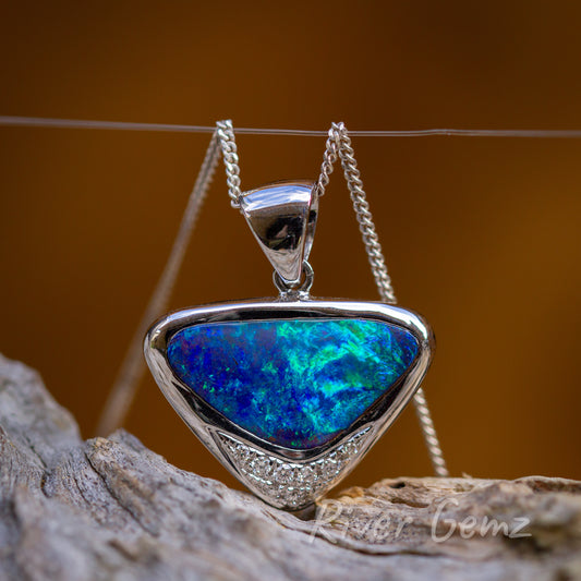 A triangular shaped blue green opal pendant with large tapered bail connected through the eyelet of the pendant. The pendant is shown against an earthy brown/orange backdrop and sits on a light grey textured piece of wood.