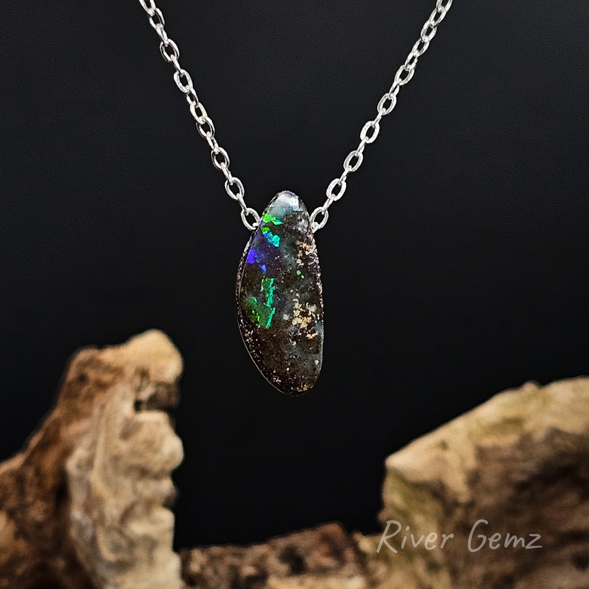 Boulder opal necklace with a silver chain against a black background and drift wood at the base with 'River Gemz' water mark in the bottom right hand corner.