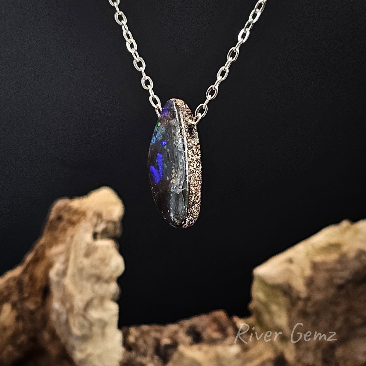 A silver chain necklace with a pendant featuring a dark, iridescent opal-like gemstone with vivid blue and purple flashes. The necklace is displayed against a dark background with decorative driftwood props at the bottom.