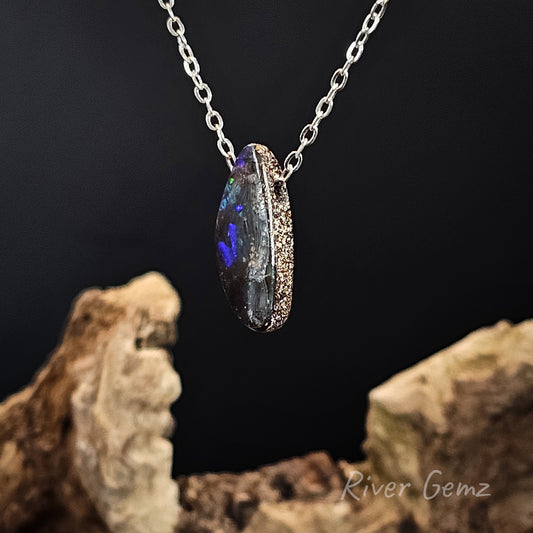 A silver chain necklace with a pendant featuring a dark, iridescent opal-like gemstone with vivid blue and purple flashes. The necklace is displayed against a dark background with decorative driftwood props at the bottom.