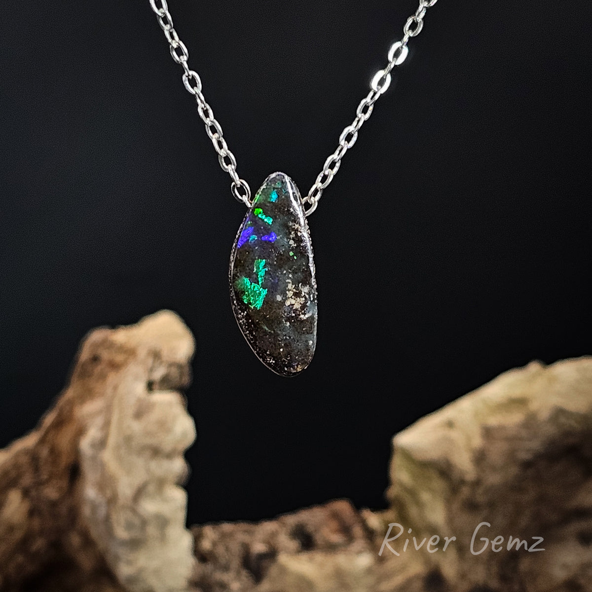 A silver chain necklace with a free-form cut opal pendant, displayed against a dark background with pieces of driftwood. The opal shows vibrant green and blue flashes.