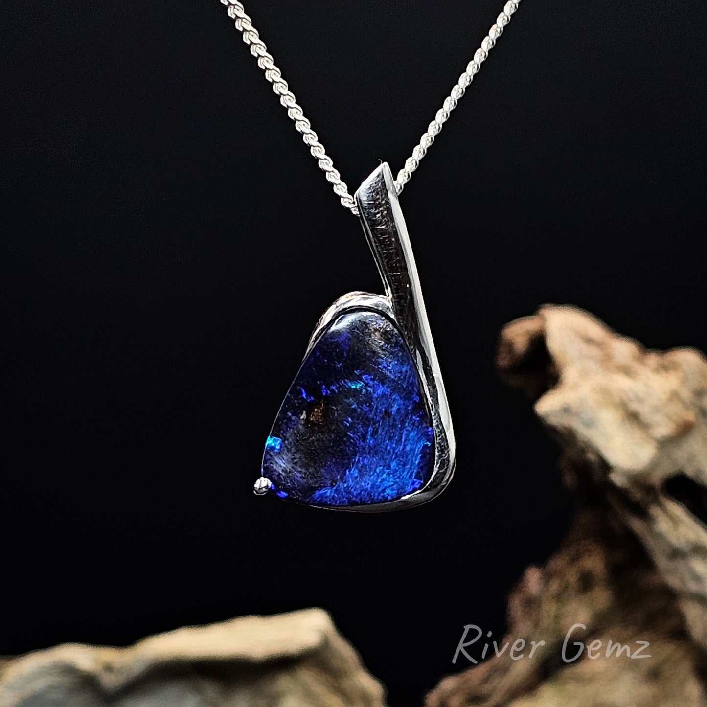 Dark blue boulder opal set in silver pendant on a dark background with 'River Gemz' branding.
