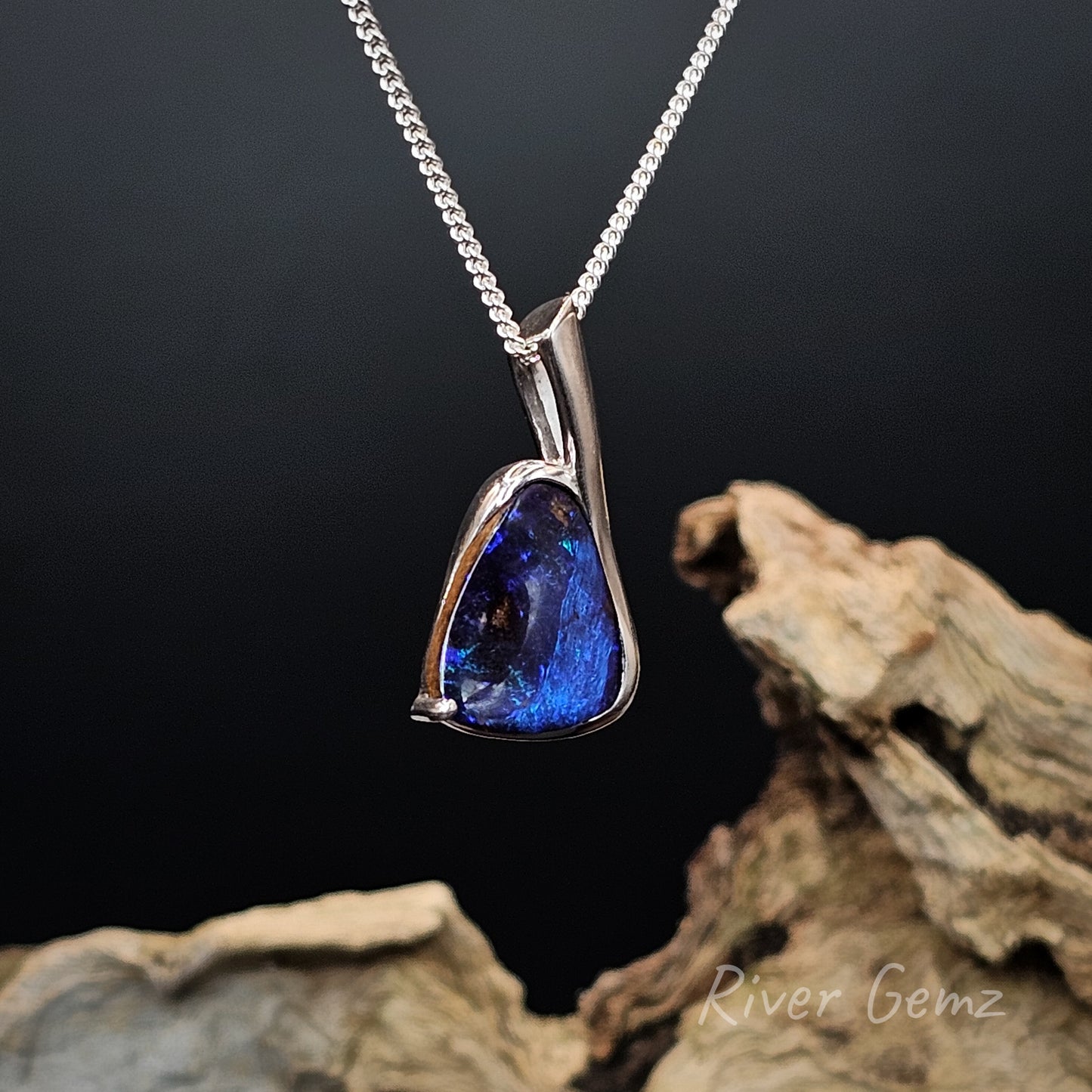 Blue pendant necklace on a chain with a dark background and 'River Gemz' branding.