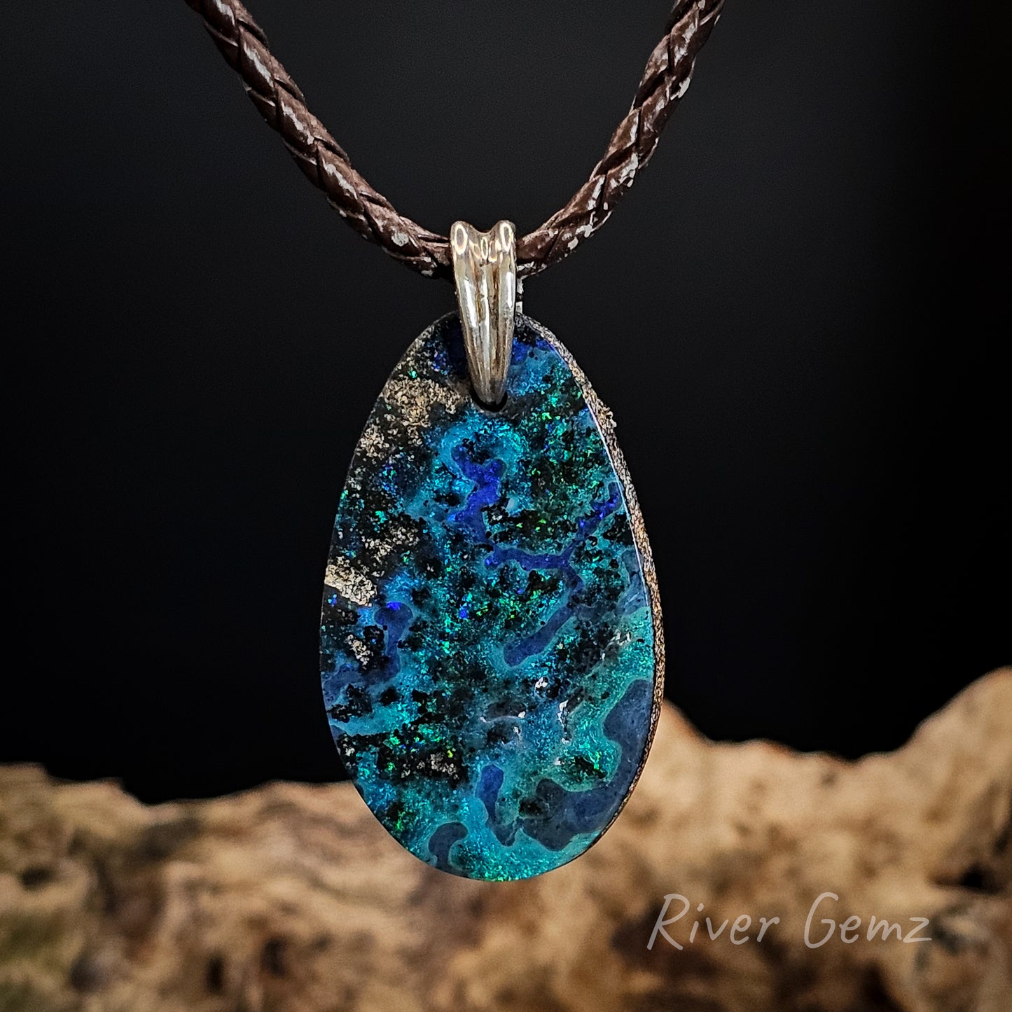Teardrop-shaped blue and green pendant on a braided brown cord against a dark background with 'River Gemz' branding.