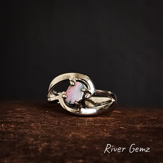 Silver ring with an oval shaped opal stone on a rusty coloured surface, branded 'River Gemz'.