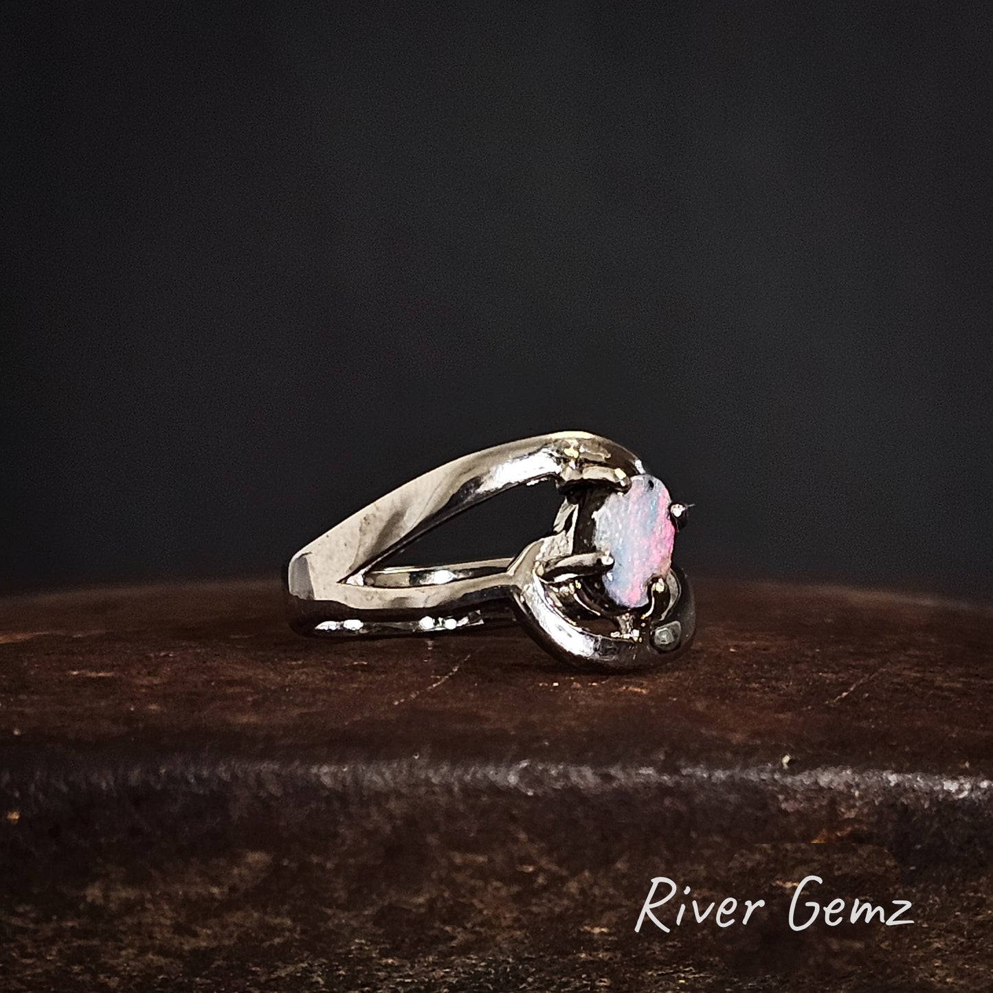 Side view of silver ring with a pink and blue opal on a dark surface, branded 'River Gemz'.