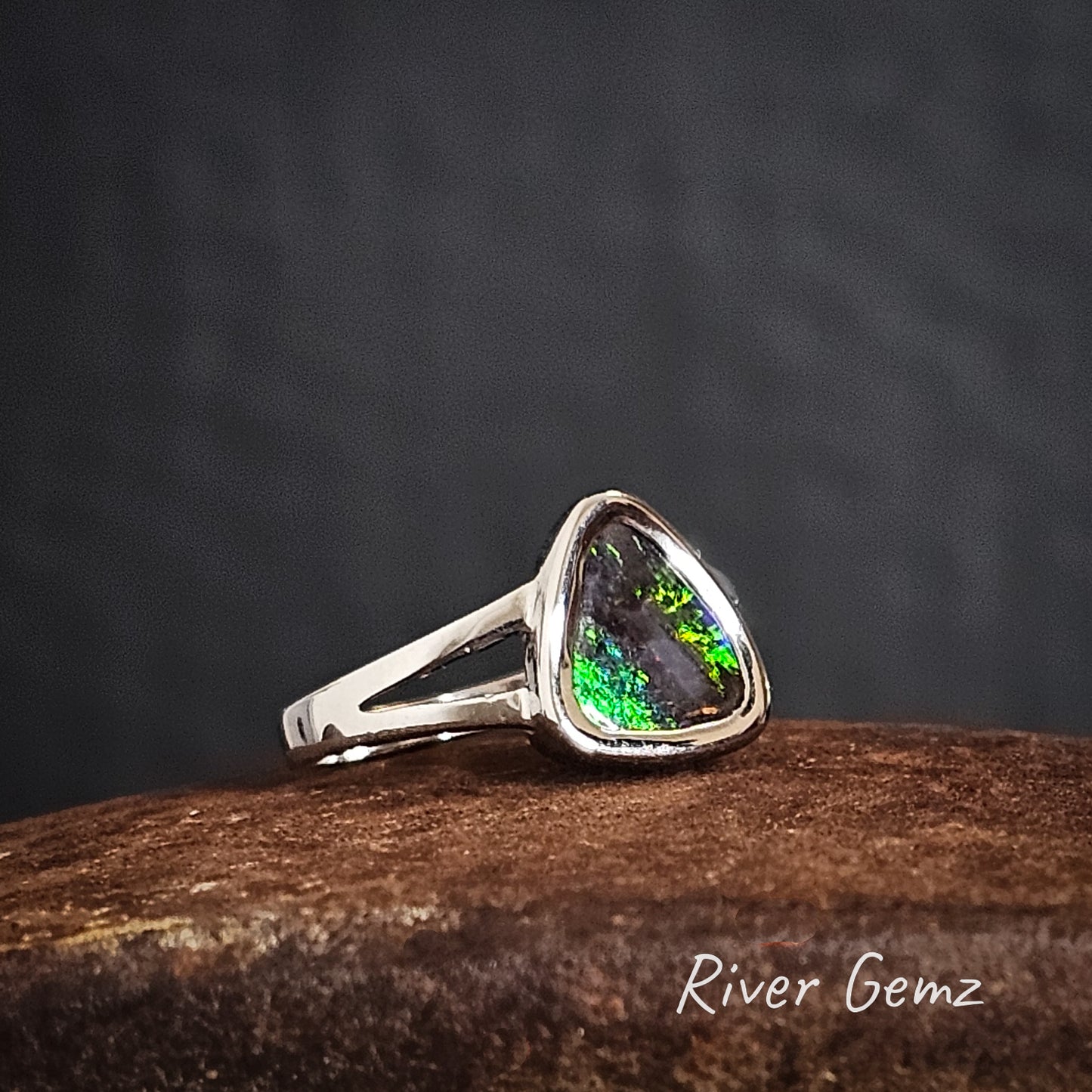Sterling silver ring with forked shoulders with a triangular shaped green and black gemstone on a dark background, branded 'River Gemz'.