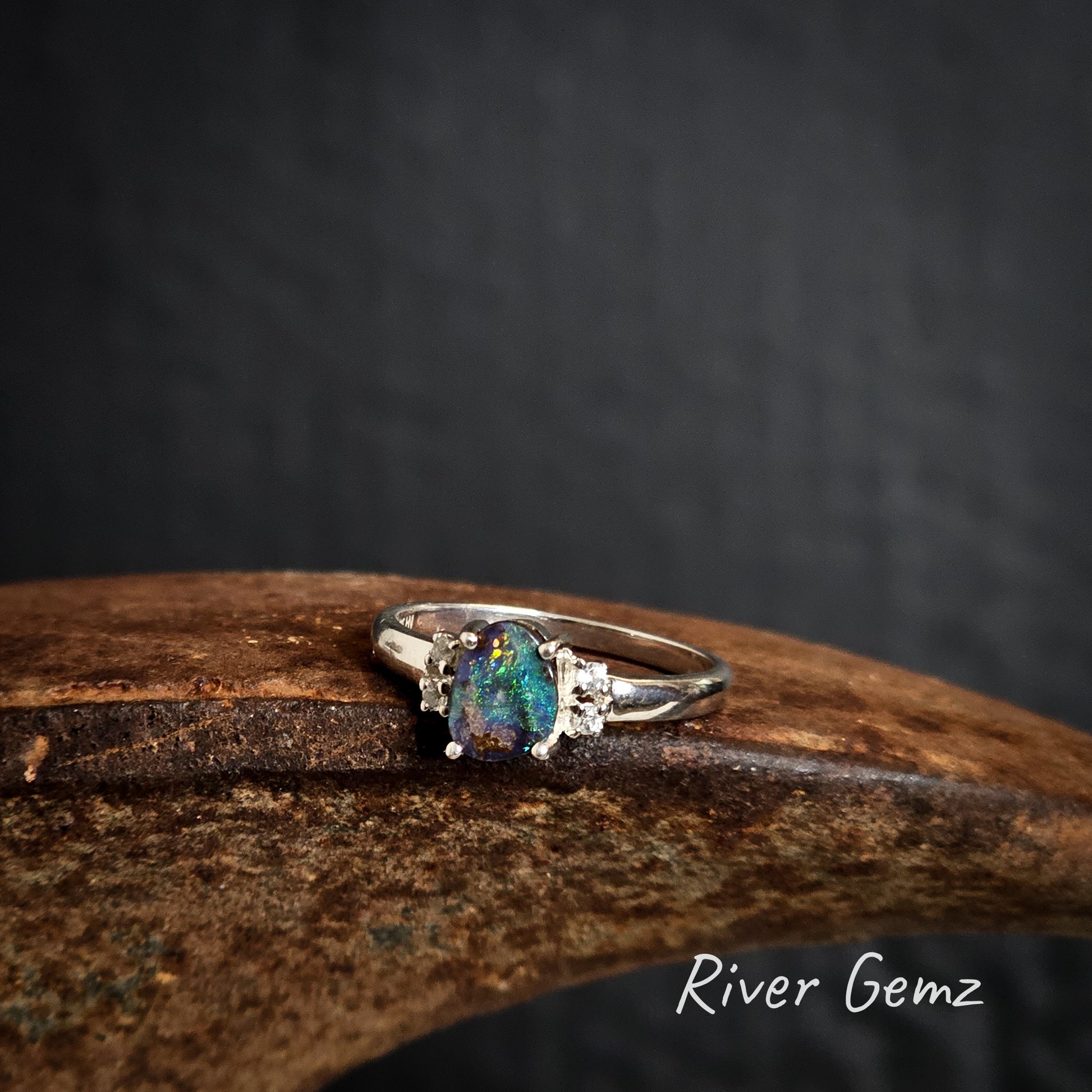 Silver ring with a green and pink boulder opal on a rusty surface, branded 'River Gemz'.